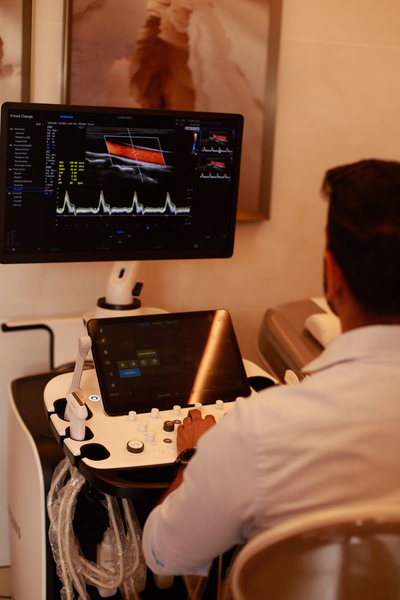 Doctor using an ultrasound machine, with a monitor displaying vascular ultrasound results in a medical examination room.